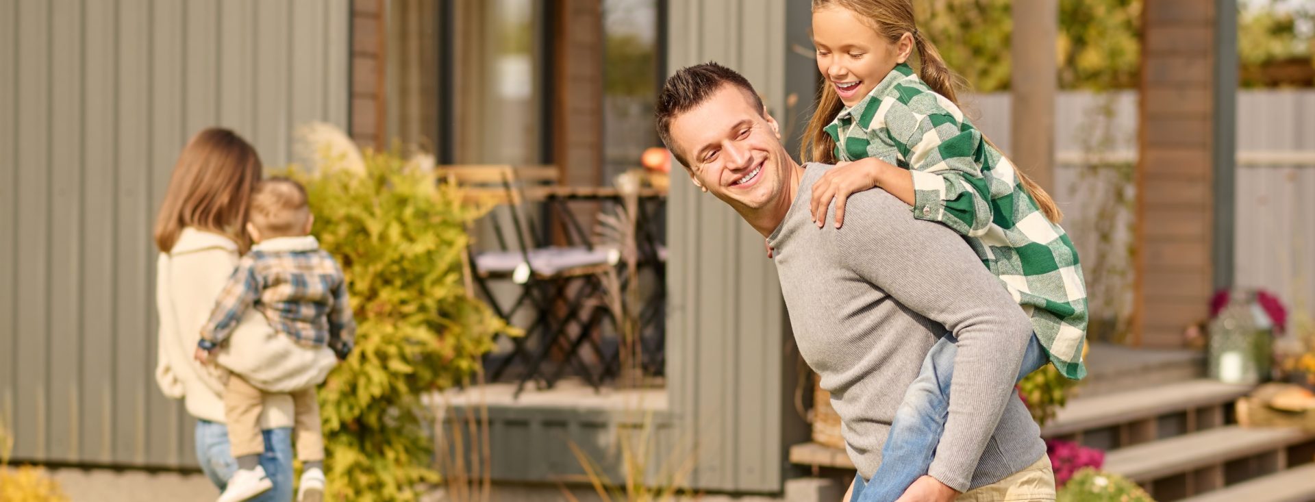 Playful mood. Smiling young adult man in casual clothes holding enthusiastic school-age daughter on back and woman with child with back to camera in courtyard of country house