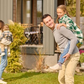 Playful mood. Smiling young adult man in casual clothes holding enthusiastic school-age daughter on back and woman with child with back to camera in courtyard of country house