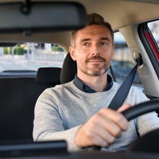 Mid adult casual man smiling while driving car and looking at mirror for reverse. Happy man feeling comfortable sitting on driver seat in his new car. Smiling mature businessman with seat belt on driving vehicle for transport and copy space.