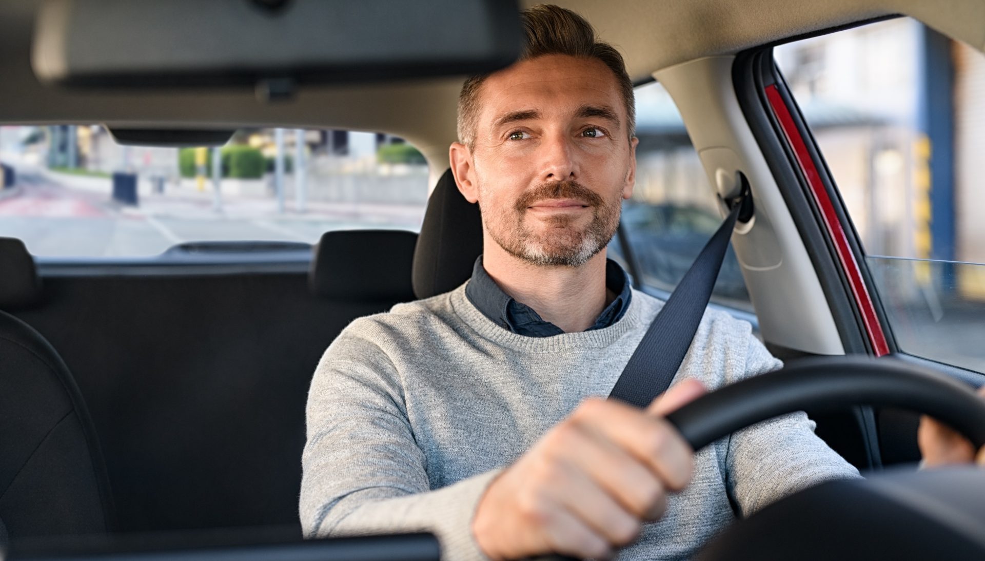 Mid adult casual man smiling while driving car and looking at mirror for reverse. Happy man feeling comfortable sitting on driver seat in his new car. Smiling mature businessman with seat belt on driving vehicle for transport and copy space.