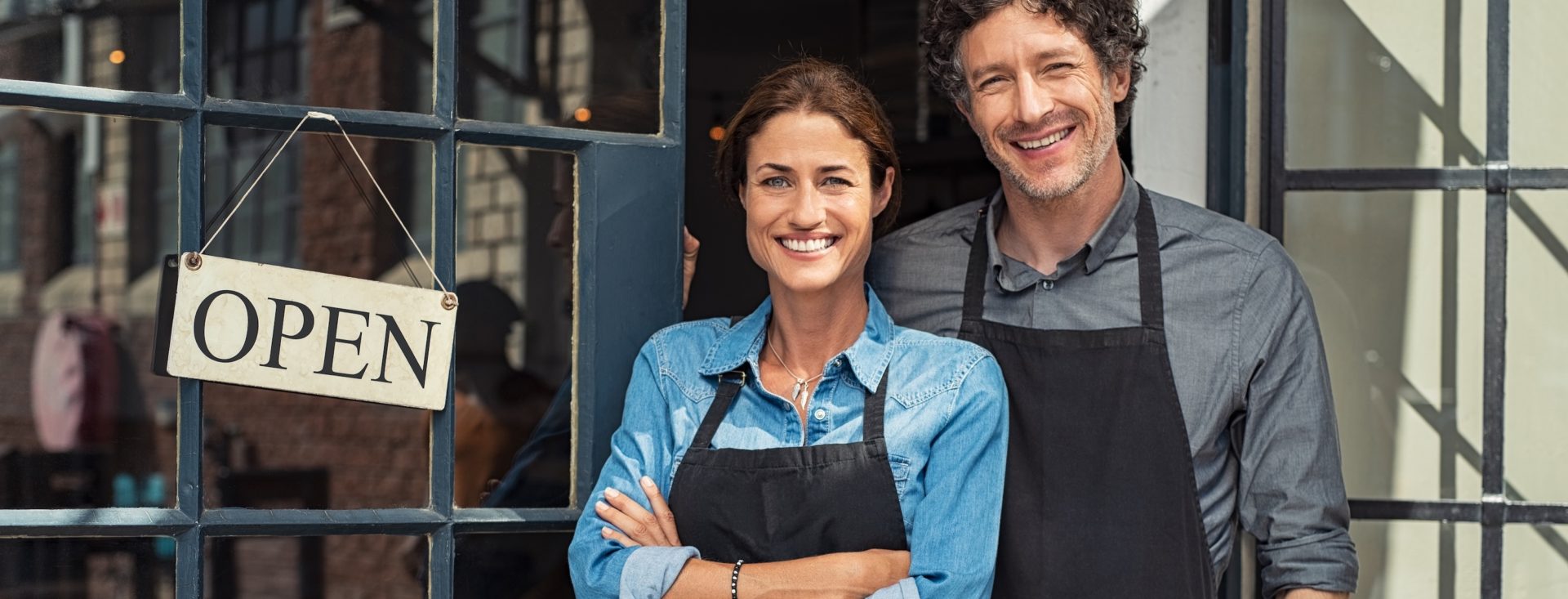 Two cheerful small business owners smiling and looking at camera while standing at entrance door. Happy mature man and mid woman at entrance of newly opened restaurant with open sign board. Smiling couple welcoming customers to small business shop.